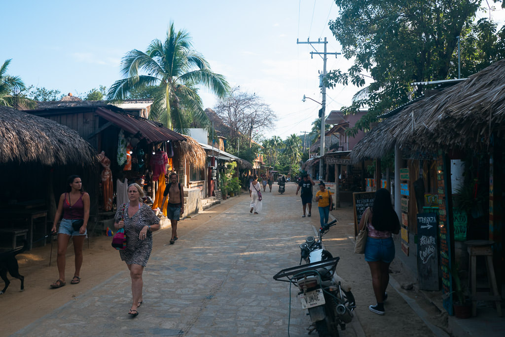 A sunny street in Mazunte, Oaxaca lined with small shops and thatched-roof buildings, with people strolling along the sandy path and a palm tree visible in the background.