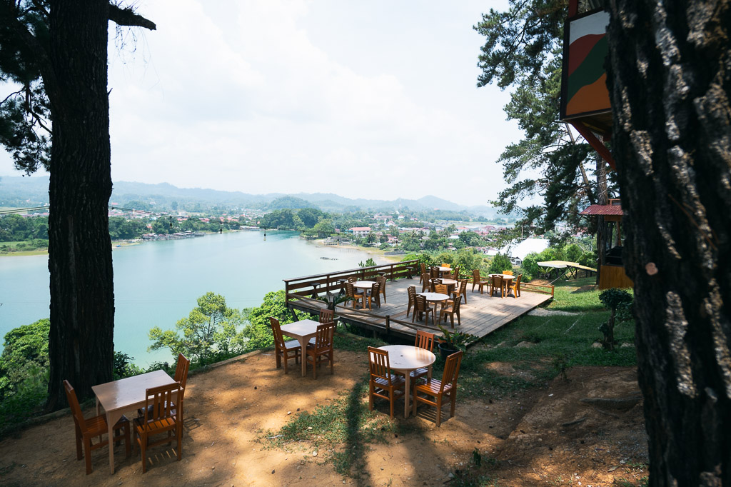 A high-angle view from the Atalambu Hill Cafe showing an outdoor seating area with several wooden tables and chairs on a dirt patch in the foreground